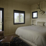 Hunting cabin bedroom interior. Walls and ceiling have wood panel finish. It has two dark framed windows and it's own outside door, leading out to the enclosed porch.