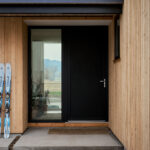 Close-up view of a modern home's entrance featuring natural wood siding and a sleek black front door with a full-height glass sidelight. A pair of blue skis and ski poles lean against the exterior wall beside the entryway. The glass reflects the surrounding landscape, while the clean lines, warm wood textures, and contemporary design create a welcoming and minimalist appearance.