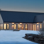 Modern single-story home with light wood siding and a black standing-seam metal roof, surrounded by snow-covered ground at dusk. Large black-framed windows and glass doors emit a warm interior glow, highlighting the home's contemporary architecture. Bare trees frame the foreground, while the soft colors of the evening sky create a peaceful rural setting.