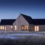 Modern contemporary home with natural wood siding and a black metal gable roof, photographed at dusk in a snowy rural landscape. Large black-framed windows and patio doors glow with warm interior light, highlighting the home's clean architectural lines. Snow-covered ground and tall grasses surround the residence, while the deep blue evening sky provides a striking backdrop.