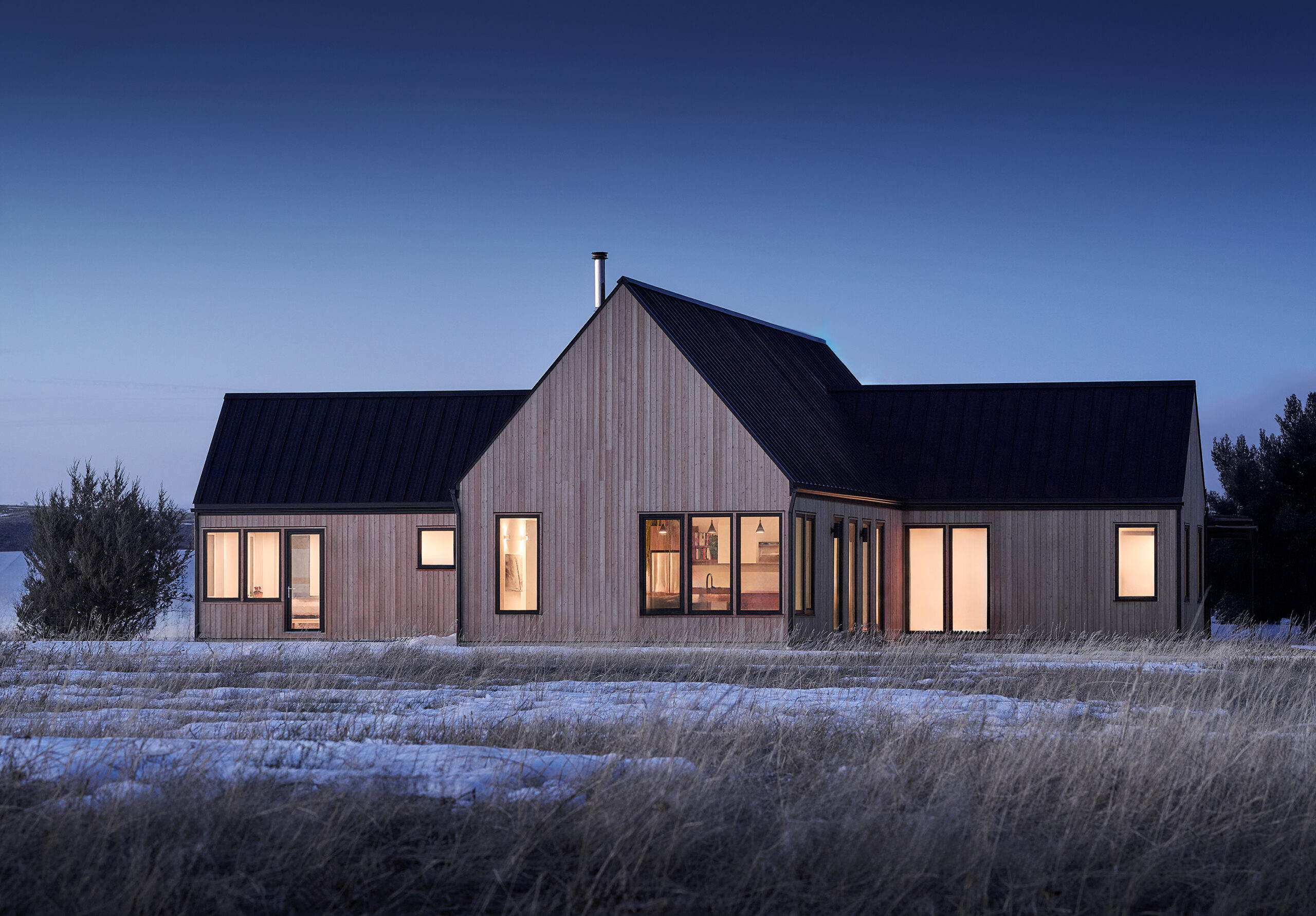 Modern contemporary home with natural wood siding and a black metal gable roof, photographed at dusk in a snowy rural landscape. Large black-framed windows and patio doors glow with warm interior light, highlighting the home's clean architectural lines. Snow-covered ground and tall grasses surround the residence, while the deep blue evening sky provides a striking backdrop.