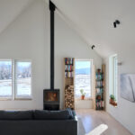 Modern living room with a vaulted ceiling, light wood floors, and large white-framed windows showcasing a snowy mountain landscape. A black wood-burning stove with a tall chimney stands between the windows, while built-in shelving filled with books, stacked firewood, and potted plants adds warmth and character. Sunlight streams into the space, illuminating a dark sofa and creating a bright, minimalist atmosphere.