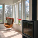 Bright modern living space featuring a corner of floor-to-ceiling white-framed windows overlooking leafless trees. Sunlight streams across light wood flooring, illuminating a tan leather lounge chair with a decorative cushion. A small vase of red tulips sits on the windowsill beside a sleek black freestanding fireplace, creating a warm and contemporary atmosphere.